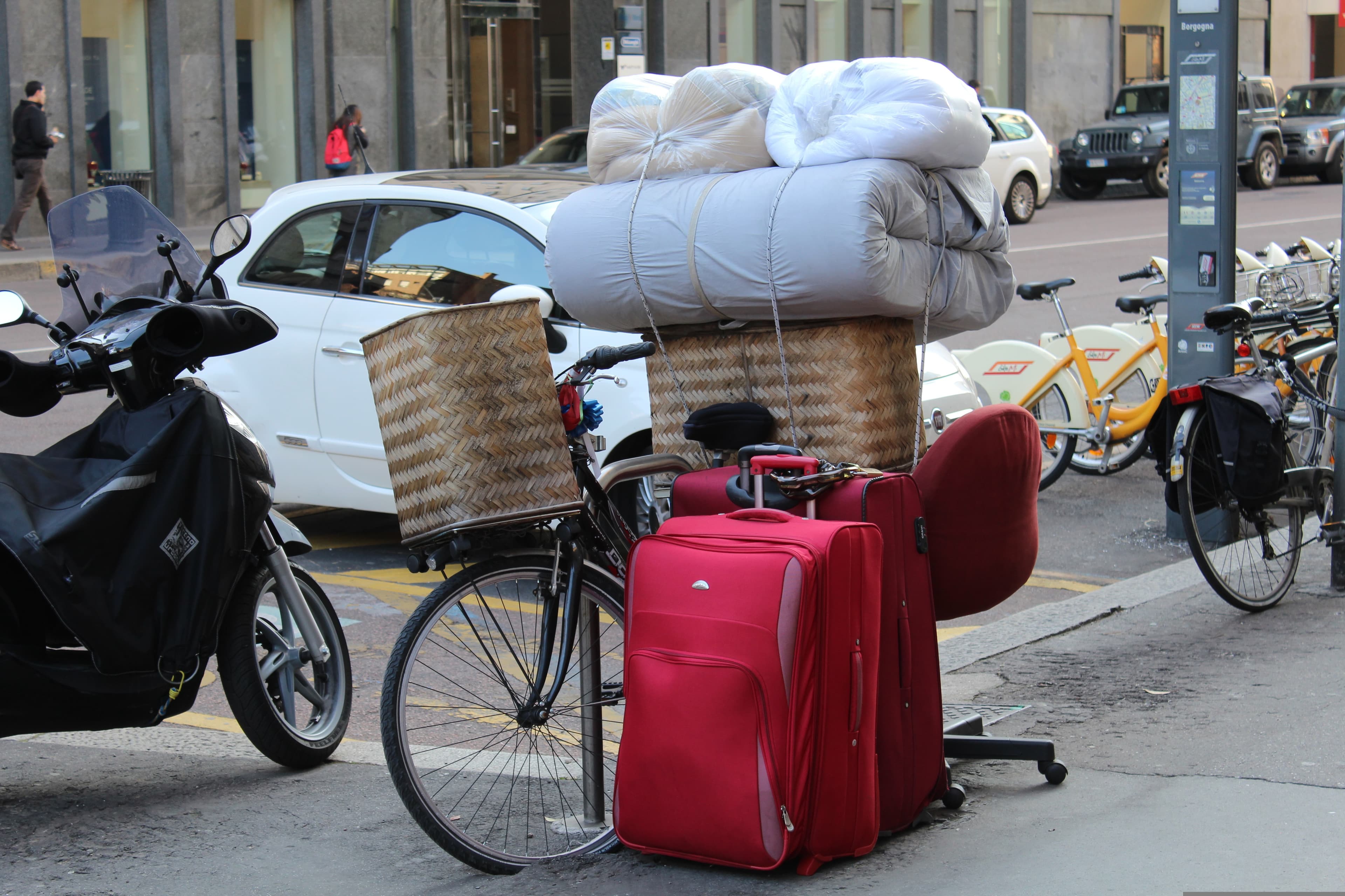 A bicycle on a city street with a mattress stacked on the front basket, a red suitcase, and an office chair beside it.