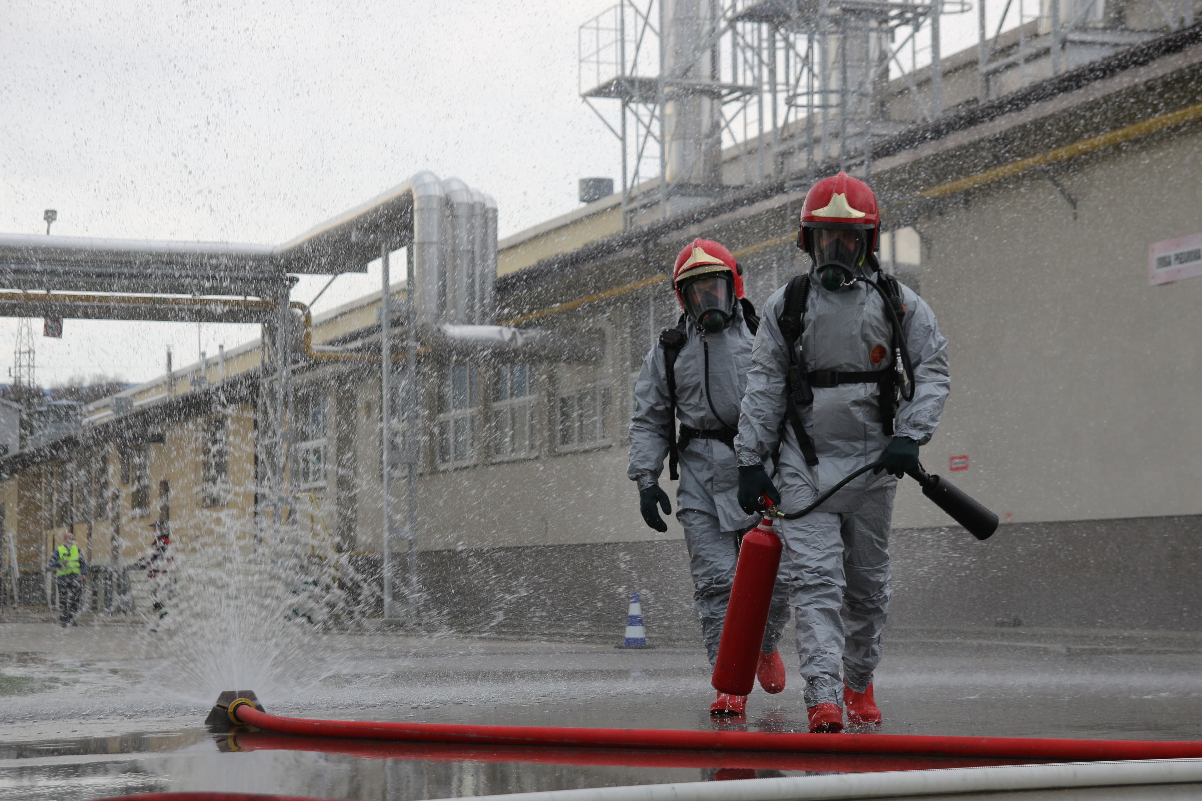 Two workers in silver fire-entry suits and breathing apparatus walking past a processing plant, a hose spraying across the yard.